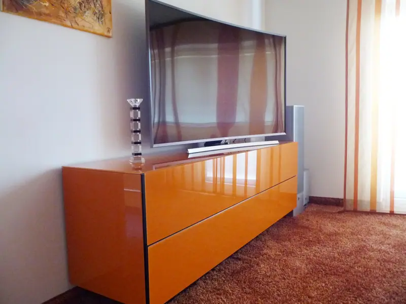 Wandhängendes Sideboard mit glänzend orangefarbenen Fronten unter einem Fernseher